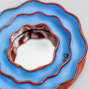 Pode incluir: Uma vista de perto de uma tigela de cer&acirc;mica com um design em camadas. A tigela apresenta um exterior azul vibrante com uma borda vermelha contrastante. O interior tem um acabamento texturizado, castanho-avermelhado. As bordas onduladas da tigela criam uma est&eacute;tica art&iacute;stica &uacute;nica.