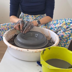 Puede incluir: Una persona est&aacute; usando un torno de alfarer&iacute;a para dar forma a un cuenco de arcilla gris. El cuenco est&aacute; sentado sobre una base de pl&aacute;stico blanca. Un cubo de pl&aacute;stico amarillo con agua de arcilla gris est&aacute; en primer plano.