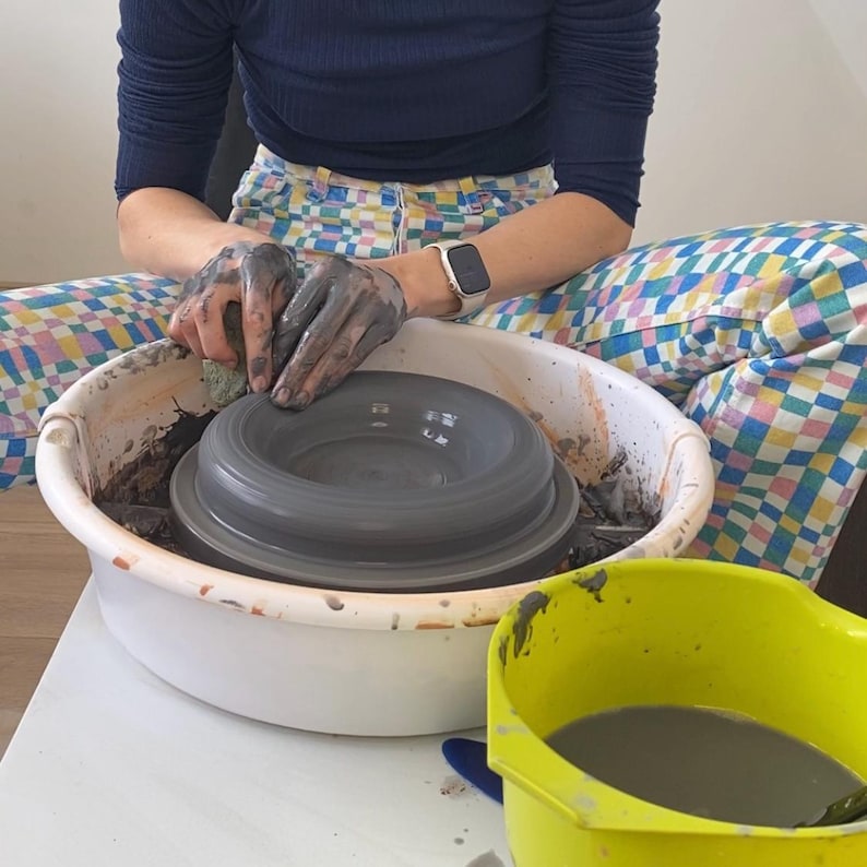 Puede incluir: Una persona est&aacute; usando un torno de alfarer&iacute;a para dar forma a un cuenco de arcilla gris. El cuenco est&aacute; sentado sobre una base de pl&aacute;stico blanca. Un cubo de pl&aacute;stico amarillo con agua de arcilla gris est&aacute; en primer plano.