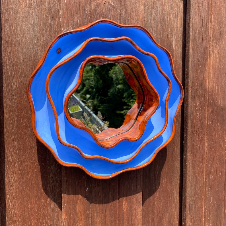 Pu&ograve; includere: Uno specchio rotondo con bordi ondulati, una cornice blu vibrante e un bordo interno marrone caldo. Lo specchio riflette una scena naturale all'aperto. Il pezzo decorativo &egrave; montato su una superficie di legno, aggiungendo un tocco artistico a qualsiasi spazio.