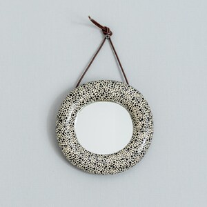 May include: Round wall mirror with a speckled black and white design. The mirror is suspended by a brown leather strap. The mirror's frame is decorated with small black dots on a white background.