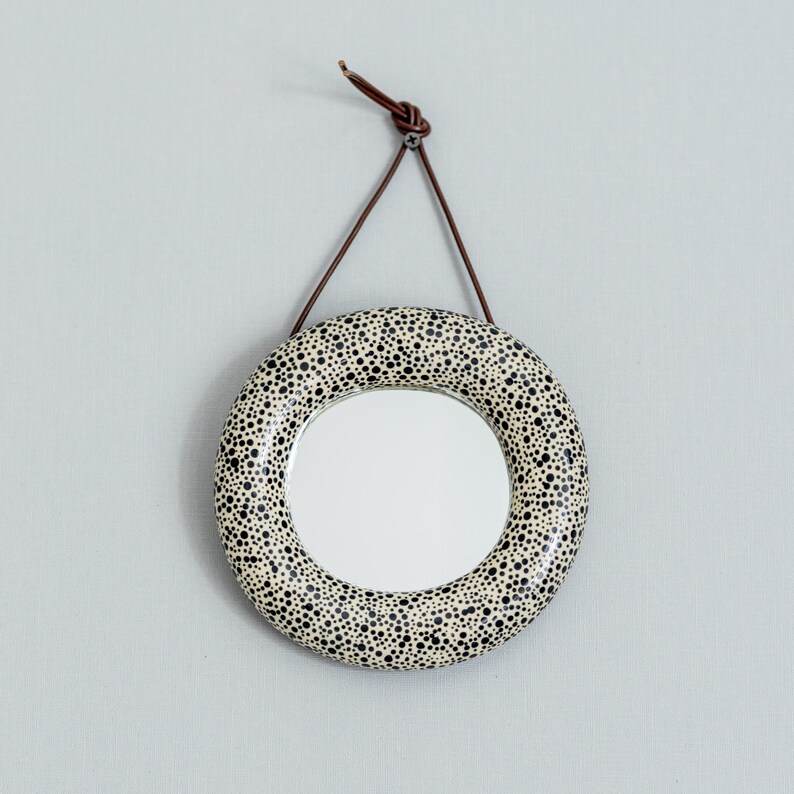 May include: Round wall mirror with a speckled black and white design. The mirror is suspended by a brown leather strap. The mirror's frame is decorated with small black dots on a white background.