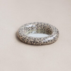 May include: A round decorative mirror with a speckled pattern. The mirror's frame is a ring shape, covered in small black dots on a cream background. The mirror itself is a reflective surface.