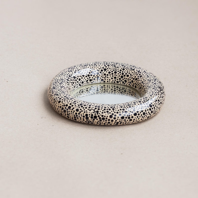 May include: A round decorative mirror with a speckled pattern. The mirror's frame is a ring shape, covered in small black dots on a cream background. The mirror itself is a reflective surface.