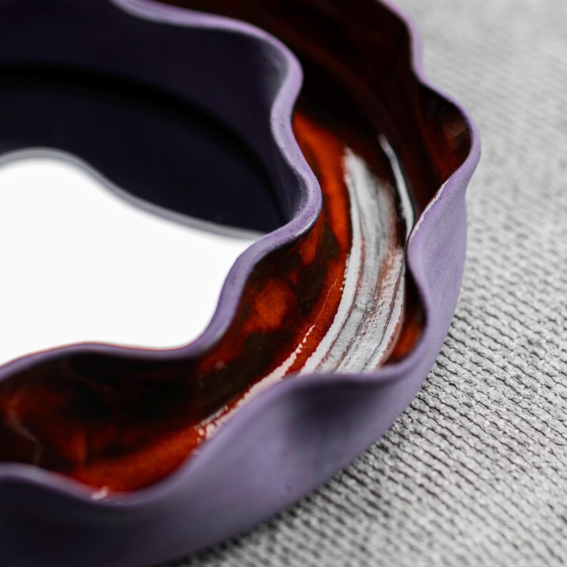 Pode incluir: Close-up de uma tigela de cer&acirc;mica feita &agrave; m&atilde;o com uma borda ondulada. A tigela apresenta um exterior roxo escuro, um interior vermelho-marrom rico e uma base interior branca. A tigela est&aacute; sobre um fundo cinza texturizado.