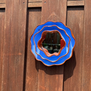 Pu&ograve; includere: Uno specchio rotondo in ceramica blu con bordo ondulato, montato su una recinzione di legno. La cornice dello specchio ha un bordo marrone a contrasto. Lo specchio riflette una vista parziale di un giardino. Un oggetto decorativo.