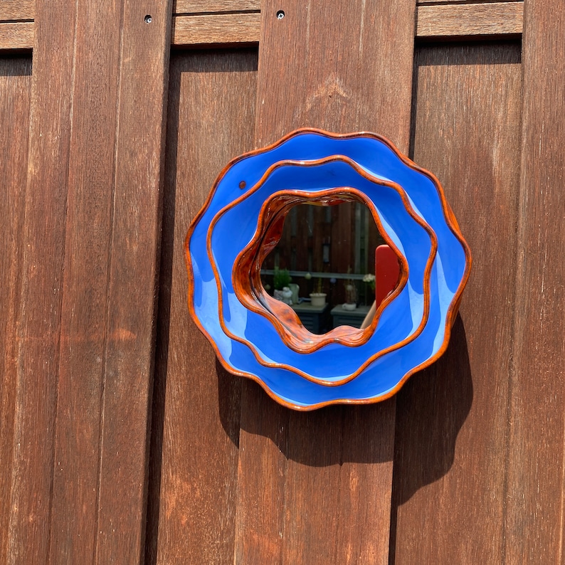 Pu&ograve; includere: Uno specchio rotondo in ceramica blu con bordo ondulato, montato su una recinzione di legno. La cornice dello specchio ha un bordo marrone a contrasto. Lo specchio riflette una vista parziale di un giardino. Un oggetto decorativo.