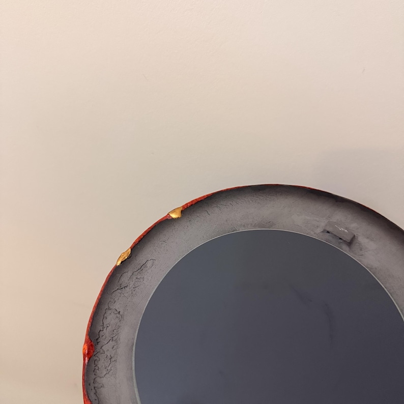 Op de afbeelding: Een ronde spiegel met een donkergrijze reflecterende oppervlakte. De lijst is een mix van rood en grijs, met gouden accenten langs de rand. De spiegel staat tegen een effen, gebroken witte muur.