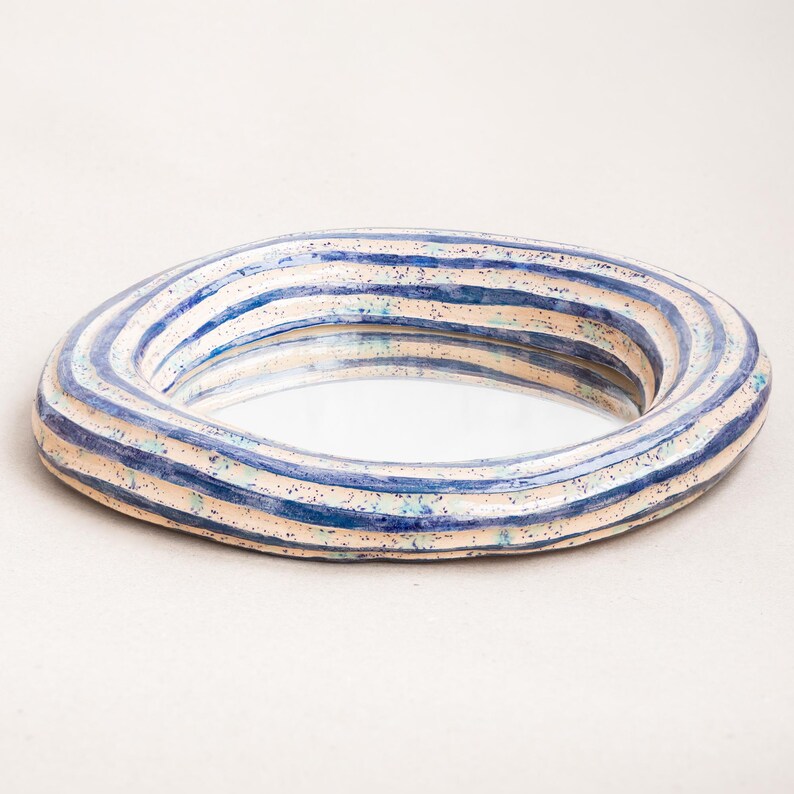 May include: A round, irregular ceramic mirror frame featuring a cream-colored base with hand-painted horizontal blue stripes and scattered dark blue and teal speckles. The frame has a thick, rounded, and textured profile surrounding a central glass mirror.