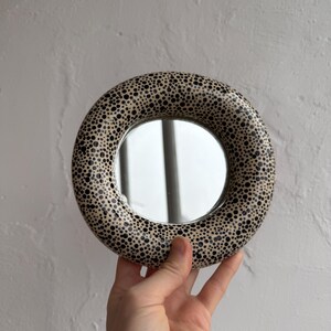 May include: A round mirror with a cream-colored frame covered in black polka dots. The mirror is held up against a white wall. The mirror's design is reminiscent of a dalmatian's coat.