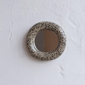 May include: A round mirror with a speckled, cream-colored frame and a black dot pattern. The mirror is mounted on a white textured wall, creating a simple yet stylish decorative piece.