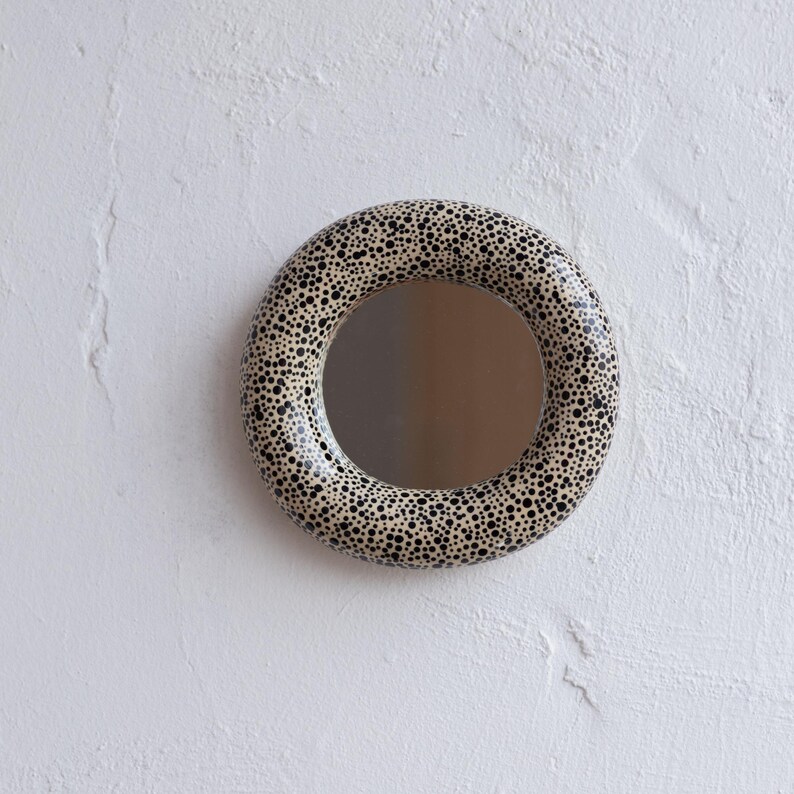 May include: A round mirror with a speckled, cream-colored frame and a black dot pattern. The mirror is mounted on a white textured wall, creating a simple yet stylish decorative piece.