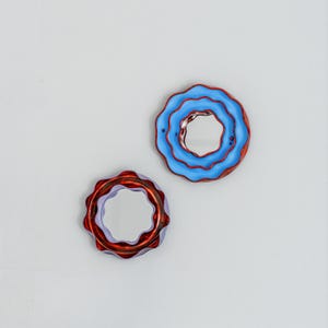 Pode incluir: Dois espelhos redondos decorativos com bordas onduladas. Um espelho &eacute; azul com borda vermelha, o outro &eacute; vermelho com borda roxa. Os espelhos s&atilde;o montados em um fundo cinza claro.
