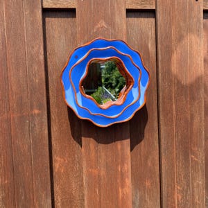 Pu&ograve; includere: Uno specchio rotondo blu con un design ondulato e stratificato. Lo specchio ha un bordo marrone ed &egrave; montato su una superficie di legno. Lo specchio riflette l'ambiente circostante.