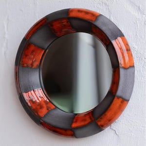 Può includere: Specchio decorativo rotondo con un motivo a scacchiera nei toni del grigio e dell'arancione. Lo specchio ha una finitura lucida ed è montato su una parete bianca. La cornice dello specchio è in ceramica.