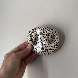 Pode incluir: Um gancho de parede de cerâmica com base oval e superfície de cor creme coberta com pontos pretos de vários tamanhos. O gancho tem duas pontas curvas que se estendem para fora e dois orifícios de montagem circulares entre as pontas.
