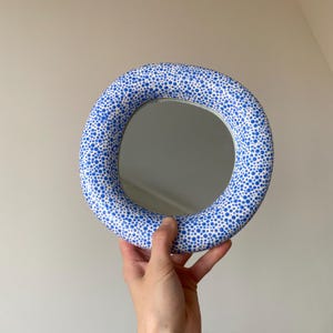 Puede incluir: Un espejo redondo con un marco blanco y lunares azules. El espejo se sostiene en una mano.