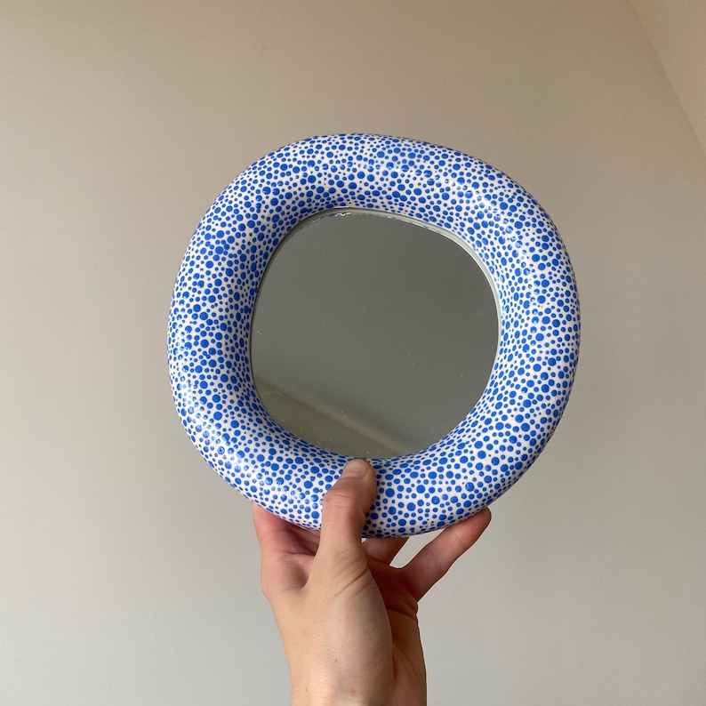 Puede incluir: Un espejo redondo con un marco blanco y lunares azules. El espejo se sostiene en una mano.