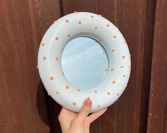 May include: A white round mirror with a raised rim and orange polka dots. The mirror is being held by a hand.