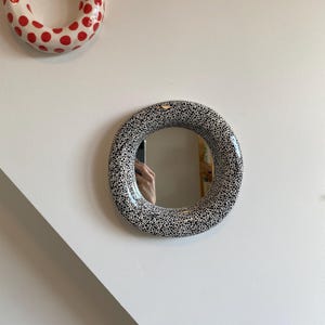 Pu&ograve; includere: Uno specchio rotondo con una cornice bianca e pois neri. Lo specchio &egrave; appeso a una parete bianca.
