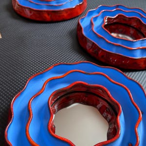 Pu&ograve; includere: Tre ciotole in ceramica con un design a strati. Le ciotole hanno un esterno blu vibrante con un bordo e un interno rossi. Le ciotole sono disposte su una superficie scura, mostrando la loro forma artistica unica.