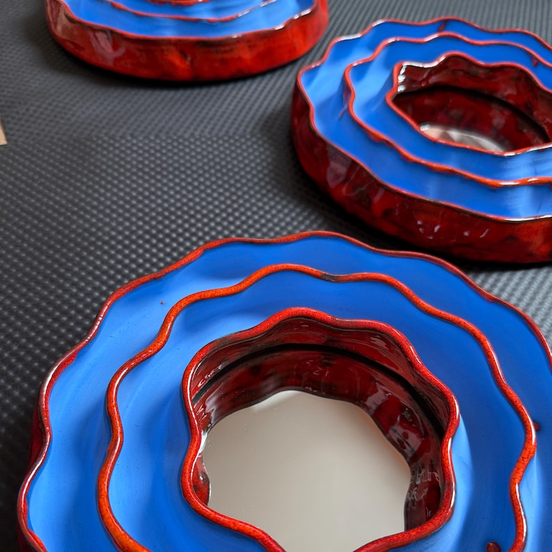 Pu&ograve; includere: Tre ciotole in ceramica con un design a strati. Le ciotole hanno un esterno blu vibrante con un bordo e un interno rossi. Le ciotole sono disposte su una superficie scura, mostrando la loro forma artistica unica.