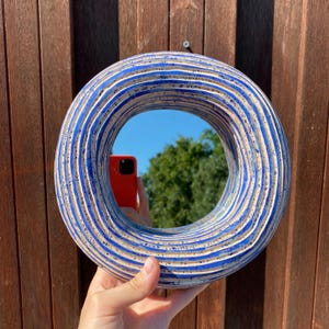 May include: A white ceramic mirror with blue and white stripes and speckles. The mirror is round and has a reflective surface.