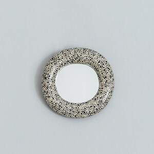 May include: Round mirror with a speckled, cream-colored frame. The frame is covered in small black dots. The mirror itself is circular and reflects a bright, white light. The mirror is a decorative item.