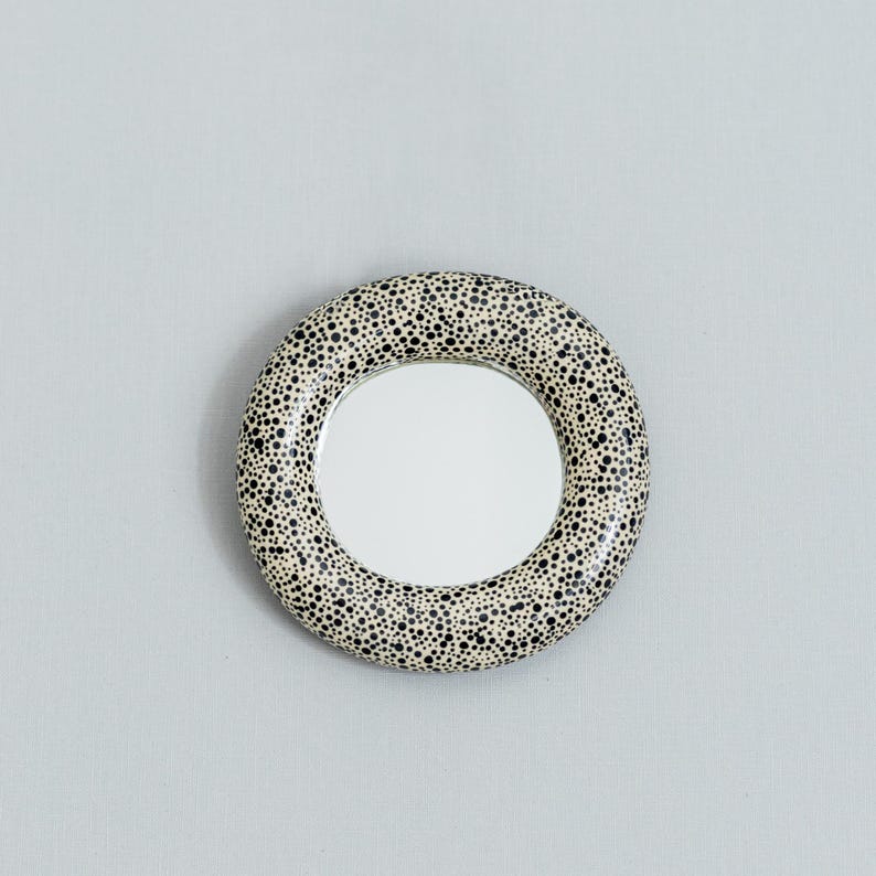 May include: Round mirror with a speckled, cream-colored frame. The frame is covered in small black dots. The mirror itself is circular and reflects a bright, white light. The mirror is a decorative item.