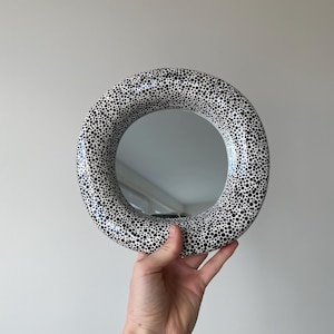 Pu&ograve; includere: Uno specchio rotondo con una cornice bianca e pois neri. Lo specchio &egrave; tenuto in mano.