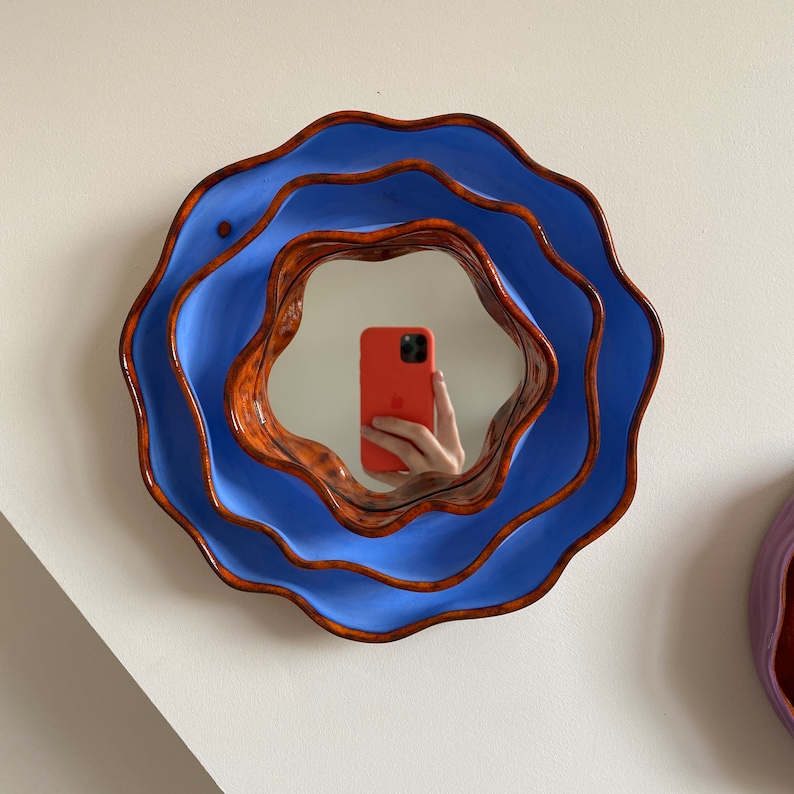 Pu&ograve; includere: Specchio da parete rotondo con un design organico a strati. Lo specchio ha una cornice esterna blu brillante e una cornice interna marrone rossiccio. Lo specchio riflette una persona che tiene in mano un telefono arancione. Lo specchio &egrave; montato su un muro bianco.