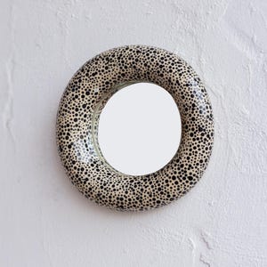 May include: A round wall mirror with a cream-colored frame covered in black polka dots. The mirror is mounted on a white textured wall, creating a striking contrast. The mirror's design adds a playful touch to any space.