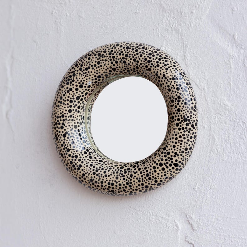 May include: A round wall mirror with a cream-colored frame covered in black polka dots. The mirror is mounted on a white textured wall, creating a striking contrast. The mirror's design adds a playful touch to any space.
