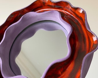 Puede incluir: Un espejo decorativo con un marco ondulado y en capas. El marco exterior es rojo vibrante con vetas marrón oscuro, mientras que el marco interior es de color lila suave. La superficie reflectante del espejo es visible.