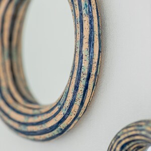 May include: A circular mirror featuring a thick, hand-crafted ceramic frame with a glossy finish. The frame displays a pattern of vertical dark blue stripes alternating with a speckled beige base, accented by subtle hints of light green throughout.