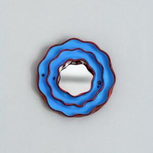 Pode incluir: Um espelho de parede decorativo redondo com um design &uacute;nico em camadas. O espelho apresenta uma moldura azul com acabamento vermelho e uma superf&iacute;cie refletora central. A moldura tem uma forma ondulada, semelhante a uma flor, adicionando um toque art&iacute;stico.