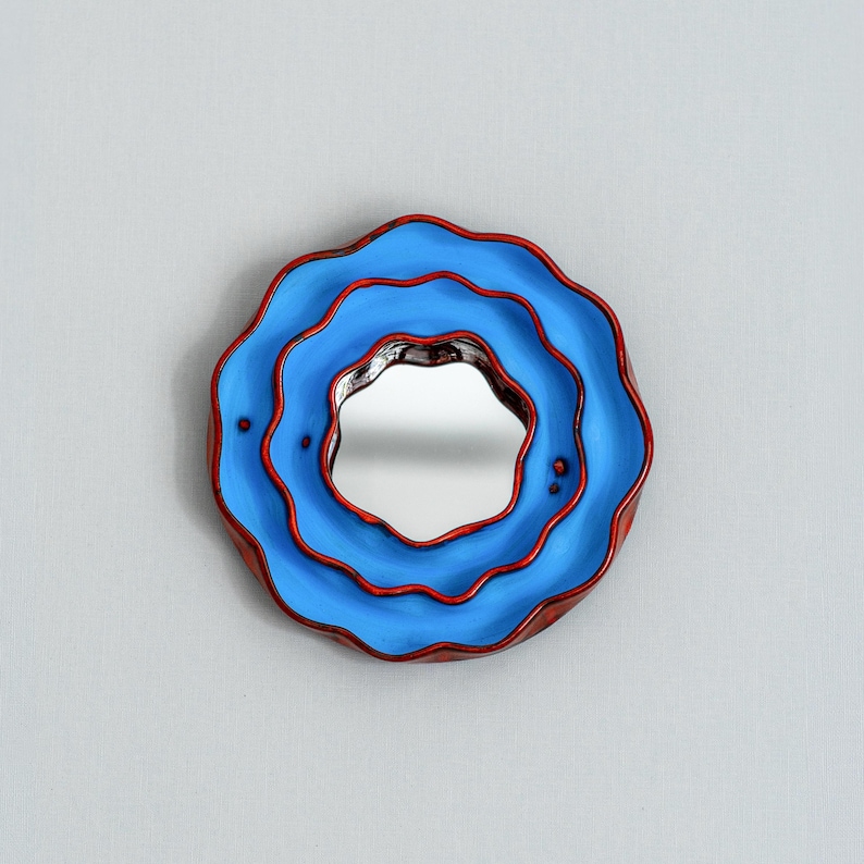 Pode incluir: Um espelho de parede decorativo redondo com um design &uacute;nico em camadas. O espelho apresenta uma moldura azul com acabamento vermelho e uma superf&iacute;cie refletora central. A moldura tem uma forma ondulada, semelhante a uma flor, adicionando um toque art&iacute;stico.