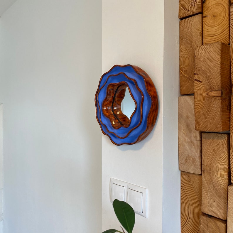 Pu&ograve; includere: Uno specchio da parete rotondo con un design unico. Lo specchio ha una cornice esterna ondulata marrone, una sezione centrale azzurra e un centro riflettente. &Egrave; montato su un muro bianco.