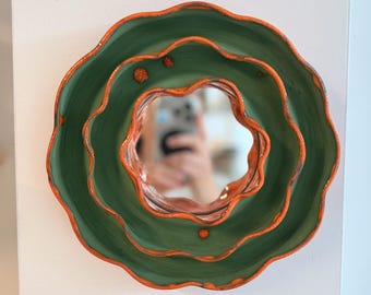 May include: A round wall mirror featuring a layered, wavy frame with a matte sage green finish and vibrant orange edges. The frame has a tiered, scalloped design with small raised orange dots on the surface, surrounding a circular reflective glass center.