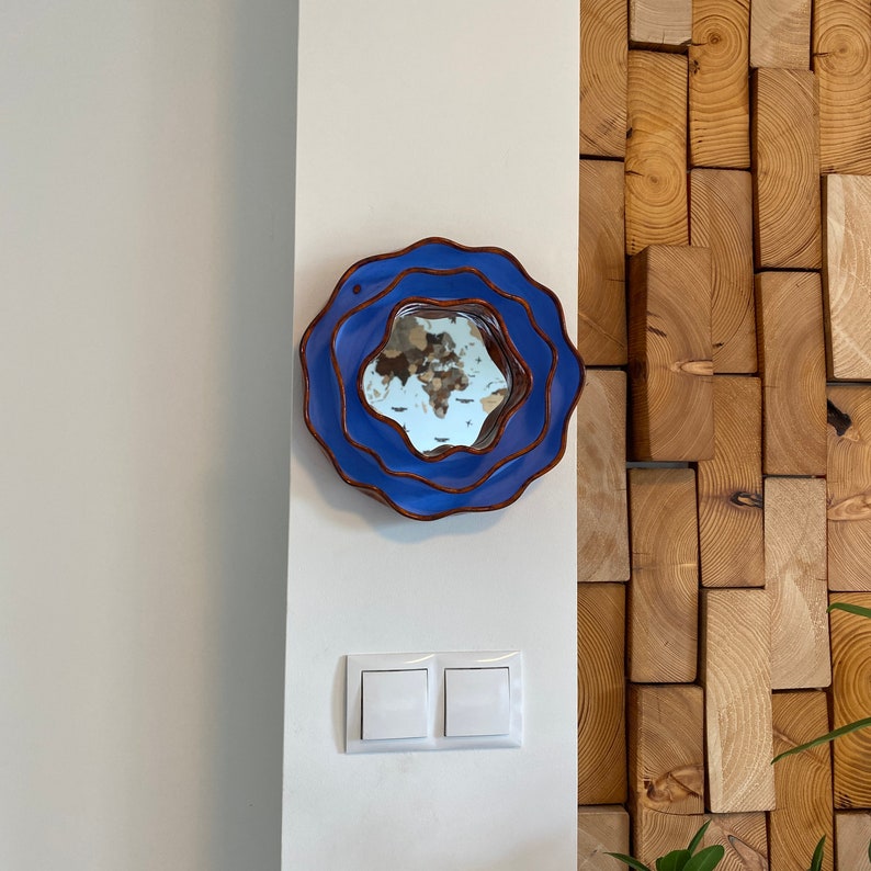 Pu&ograve; includere: Uno specchio da parete rotondo blu con un design ondulato e stratificato. Lo specchio ha un bordo marrone ed &egrave; montato su una parete bianca. La superficie riflettente dello specchio mostra una visione distorta dell'ambiente.