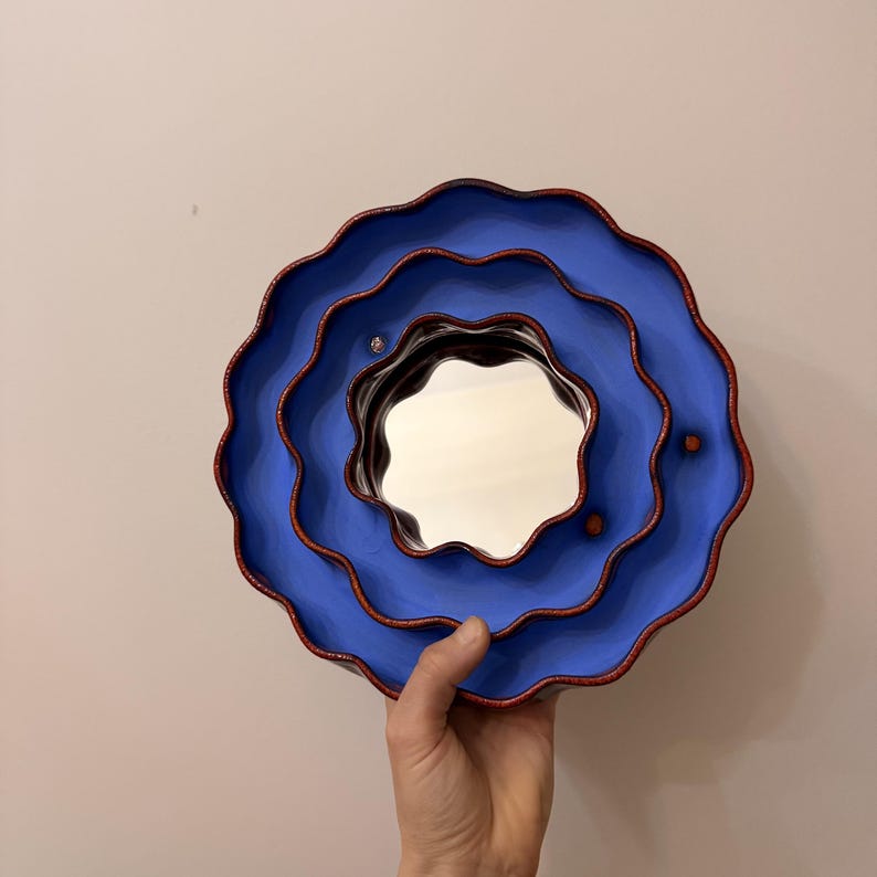 Op de afbeelding: Een unieke, gelaagde wandspiegel met een golvend, bloemachtig ontwerp. De spiegel heeft een levendig blauwe buitenkant met een rode rand en een reflecterend midden. De totale diameter is ongeveer 30 cm.