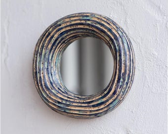 May include: Round wall mirror with a textured, ridged frame. The frame features a blend of blue, beige, and speckled accents. The mirror is mounted on a white textured wall, creating a decorative accent piece.