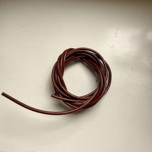 May include: A coil of dark brown leather cord, possibly for crafts or jewellery. The cord is tightly wound, with a single strand extending outwards. The leather appears smooth and has a consistent colour throughout.