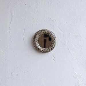 May include: A round mirror with a speckled, ring-shaped frame. The frame features a pattern of small black dots on a beige background. The mirror is mounted on a white textured wall, creating a simple, modern aesthetic.