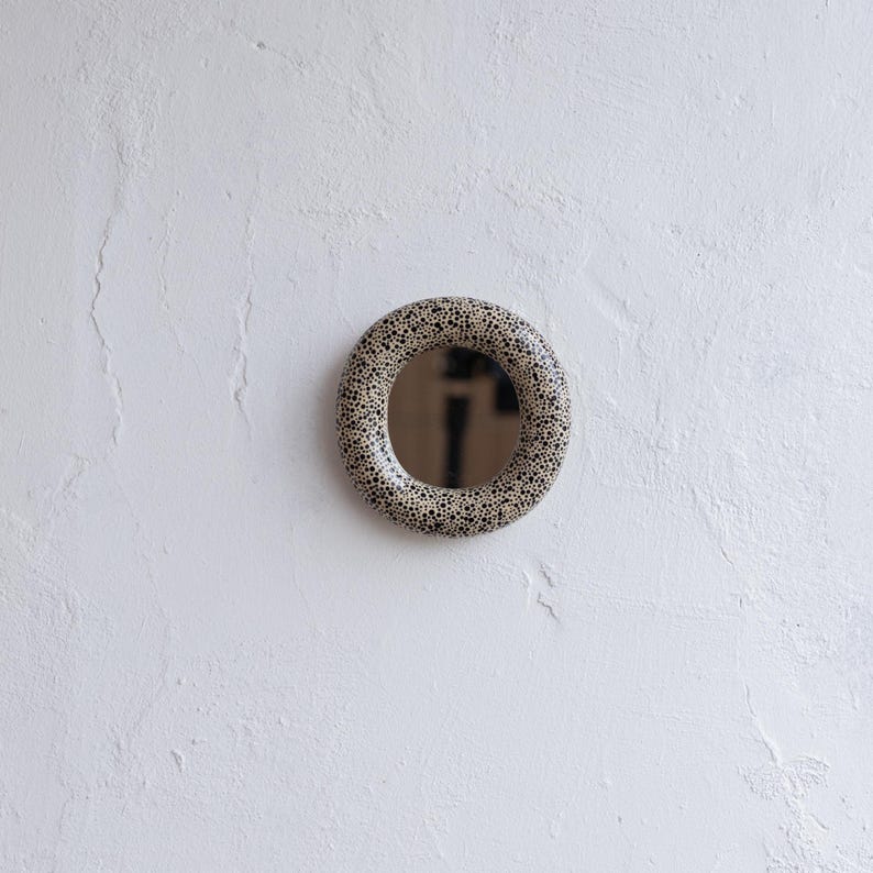 May include: A round mirror with a speckled, ring-shaped frame. The frame features a pattern of small black dots on a beige background. The mirror is mounted on a white textured wall, creating a simple, modern aesthetic.