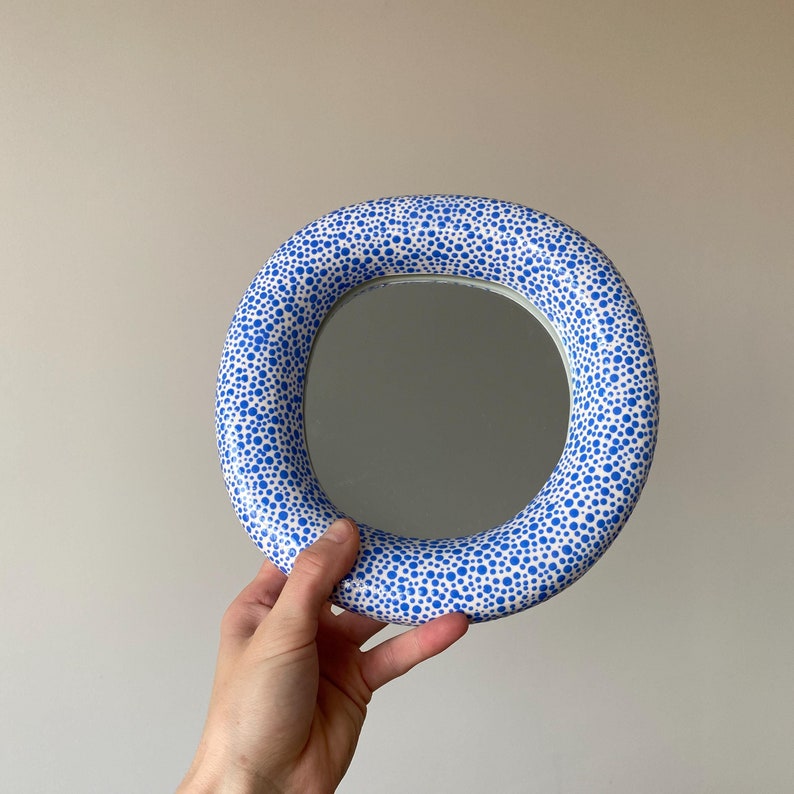 Puede incluir: Un espejo redondo con un marco blanco y lunares azules. El espejo se sostiene en una mano.