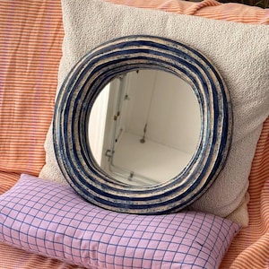 May include: A round mirror featuring a thick, textured frame with alternating concentric rings of deep navy blue and off-white glaze. The frame has a wavy, undulating surface profile, and the central glass provides a clear, reflective surface.