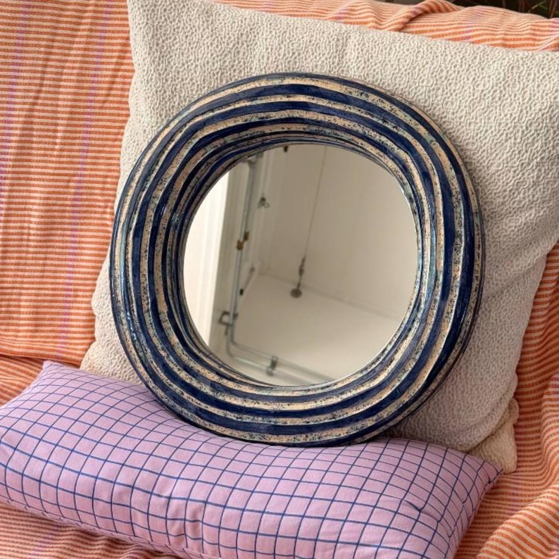 May include: A round mirror featuring a thick, textured frame with alternating concentric rings of deep navy blue and off-white glaze. The frame has a wavy, undulating surface profile, and the central glass provides a clear, reflective surface.