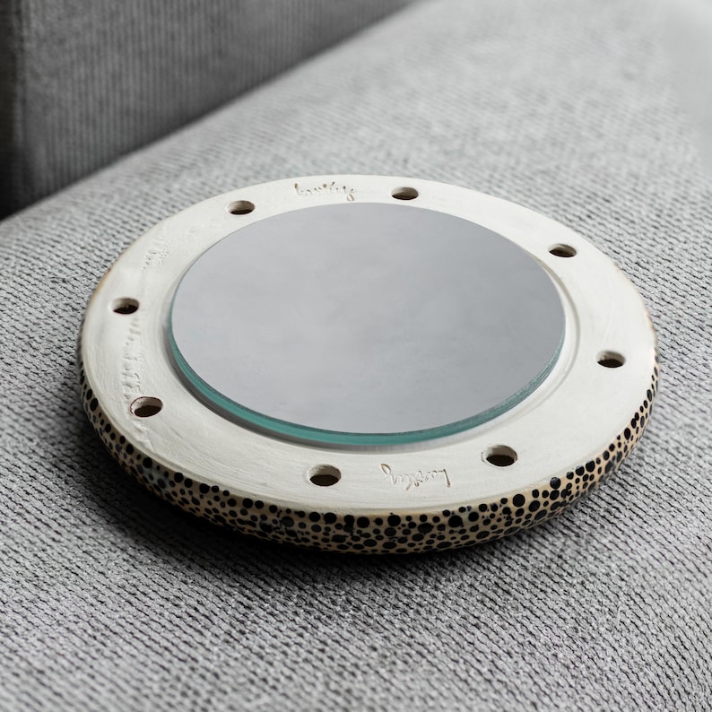 May include: A round, off-white decorative mirror with a speckled rim and a central glass mirror. The mirror has several small holes around the edge and a handwritten inscription. The mirror is resting on a gray surface.