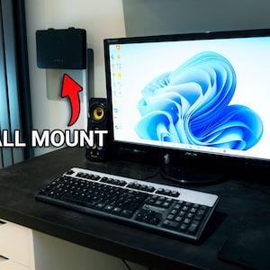 May include: A black desktop computer with a blue and white abstract background on the screen. The computer is on a black desk with a keyboard, mouse, and two black speakers. The text "WALL MOUNT" is written on the left side of the image.
